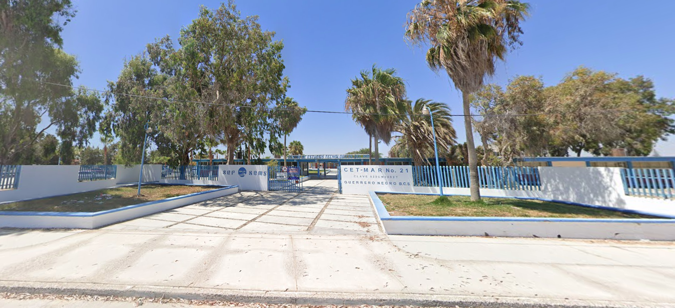 Fotografía en la que se muestra una explanada con dos jardineras grandes. Al terminar la explanada se encuentra la entrada del plantel conformada por muros blancos y rejas azules. En uno de los muros en letras grandes azules está escrito CETMAR NÚMERO 21.